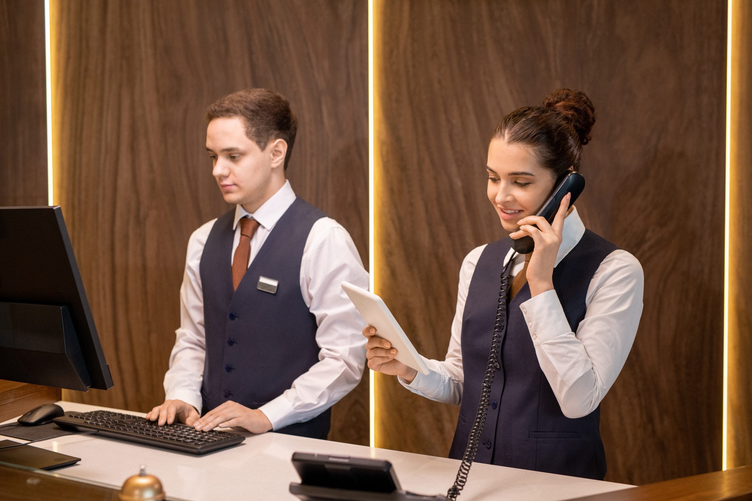 One of two young hotel receptionists standing by counter, looking at touchpad display and consulting client on the phone against colleague
